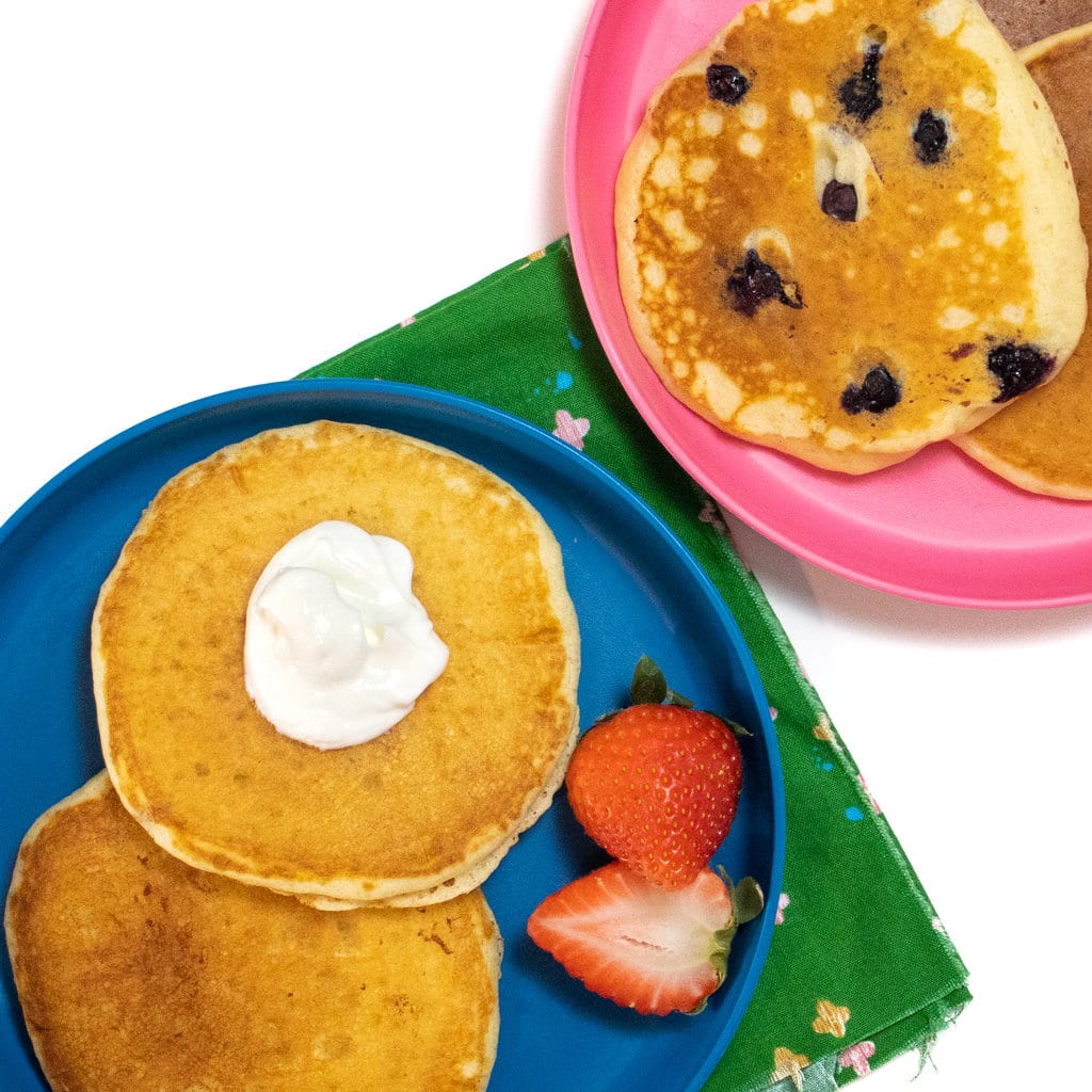 A blue and pink kids plate with yogurt pancakes on top some with blueberries on them and a side of strawberries.