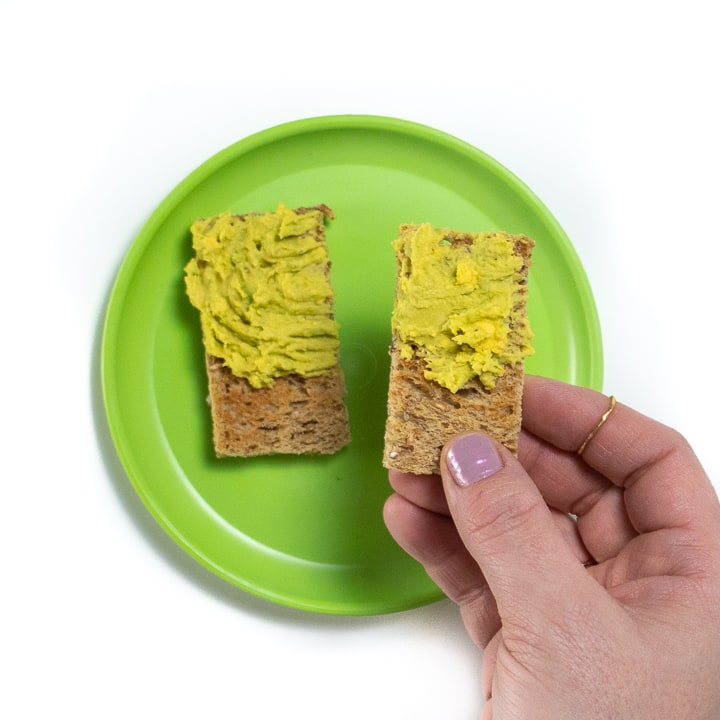 Hand holding a piece of toast with avocado and egg mashed together.