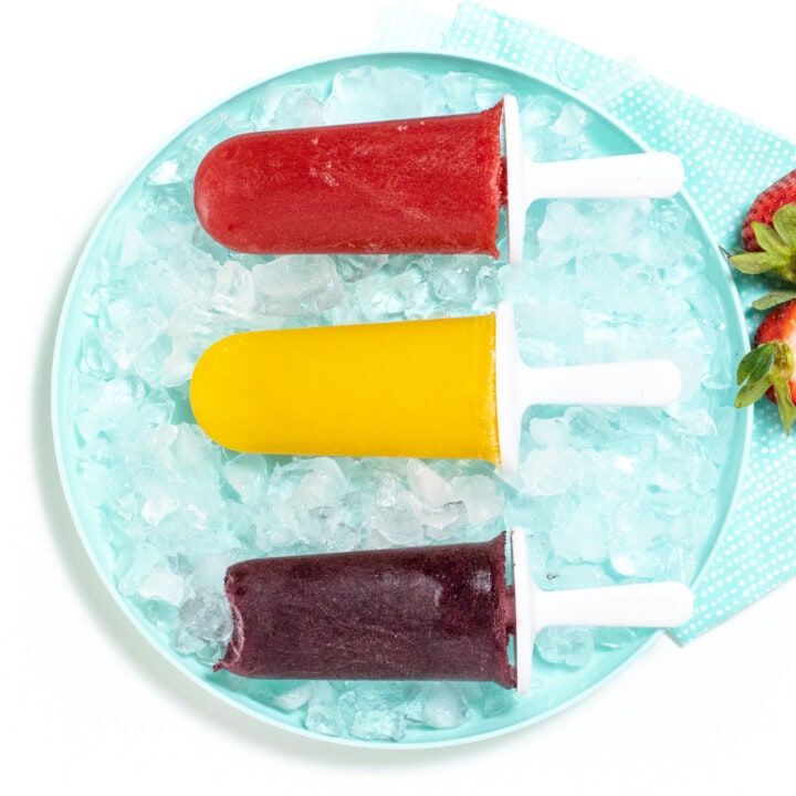 3 different fruit popsicles lined up on a blue plate with ice and a blue napkin.