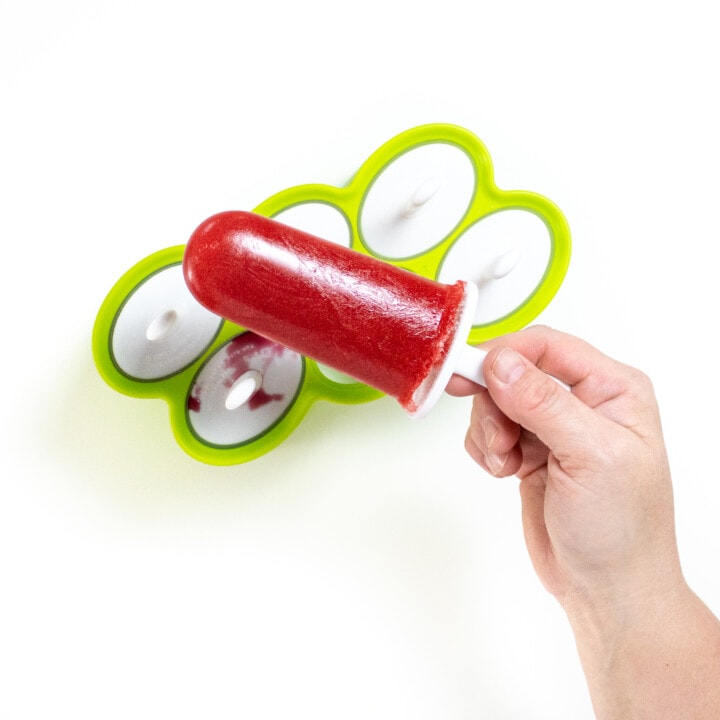 Hand holding a bright pink strawberry popsicle over a green popsicle mold.