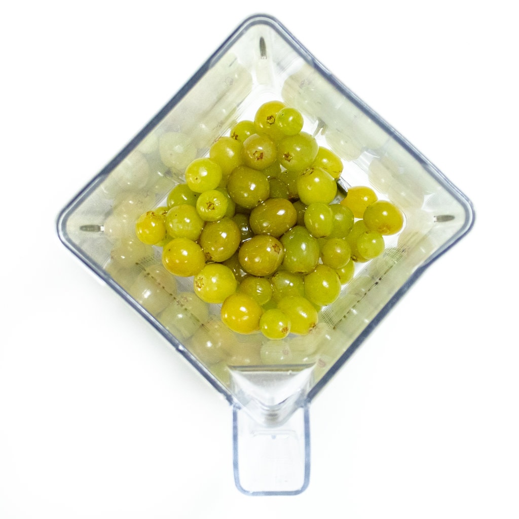 Clear blender on a white countertop with grapes, honey and grape juice inside.