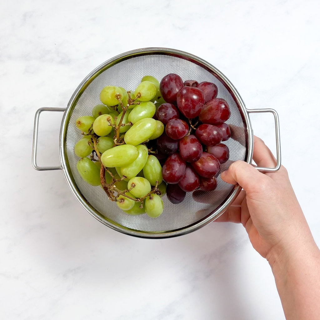 A handholding, a fine mesh colander with washed green and purple grapes over a white counter in my kitchen.