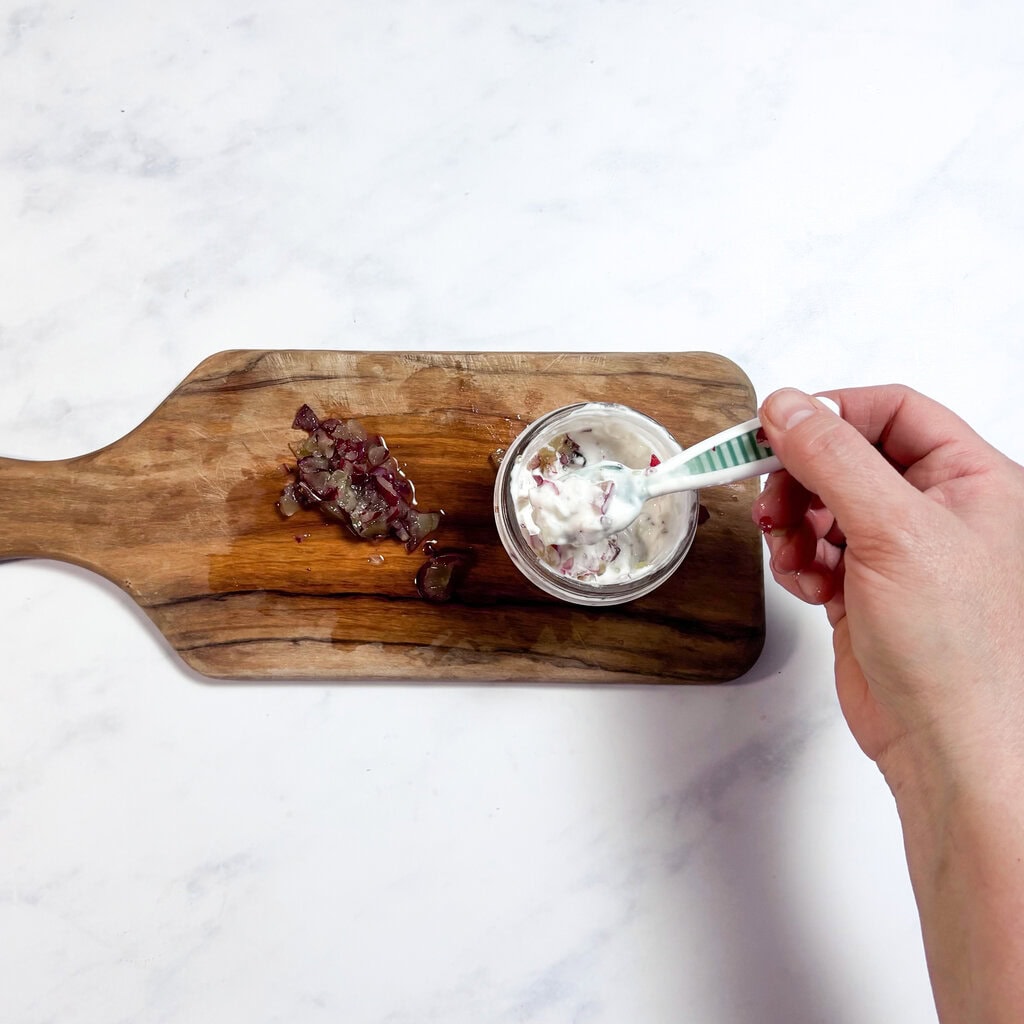 A wood cutting board on my kitchen counter with a hand, stirring a yogurt, grape purée and a small glass jar.