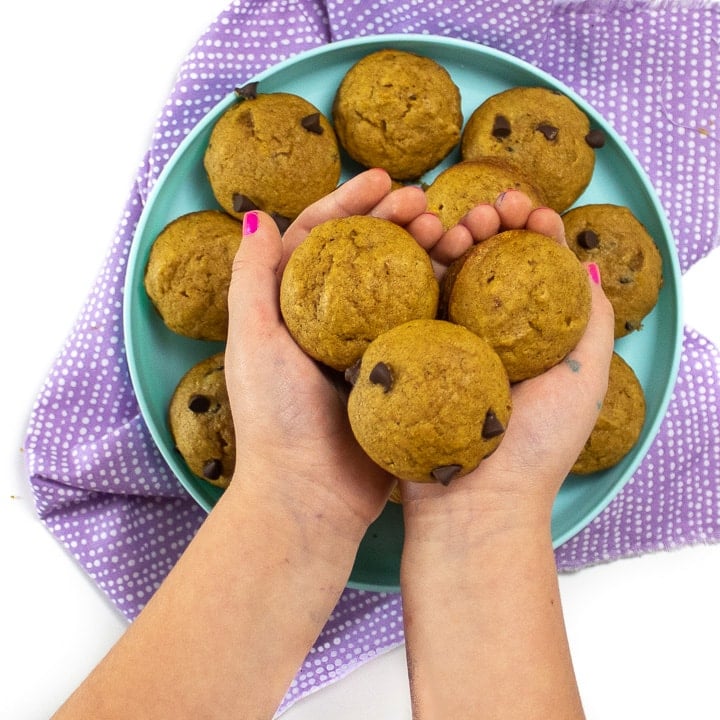 Small hands holding mini muffins in front of a teal plate with more muffins.