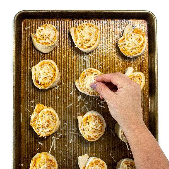 pizza dough is rolled up and sliced and on a baking sheet. Hand is sprinkling parmesan on top.