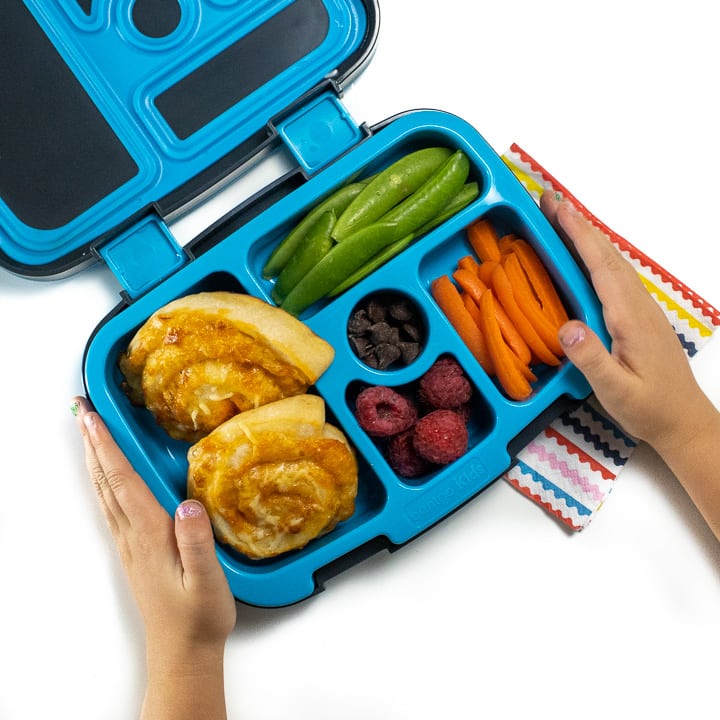 Small kids hands holding a lunch box with pizza rollups inside.