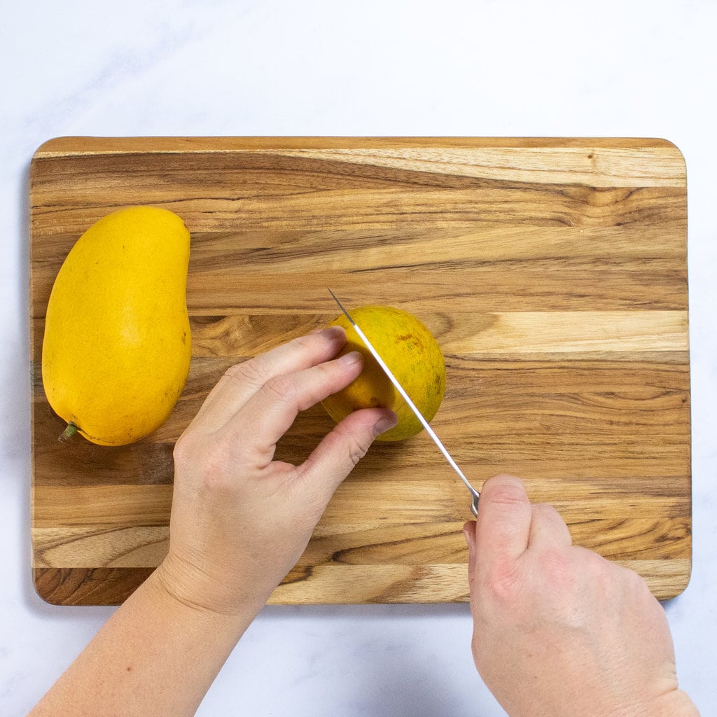 Someone cutting a mango of the pit on a wooden cutting board. Someone Someone