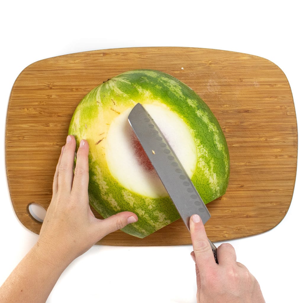 A wooden cutting board with a watermelon on its side with a hand trimming the rind off of the watermelon.