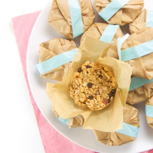 Round white plate sitting on a pink napkin, the plate is piled hight with round brown packages with blue tape holding them together. There is one package opened and there is a soft and chewy granola bar inside.