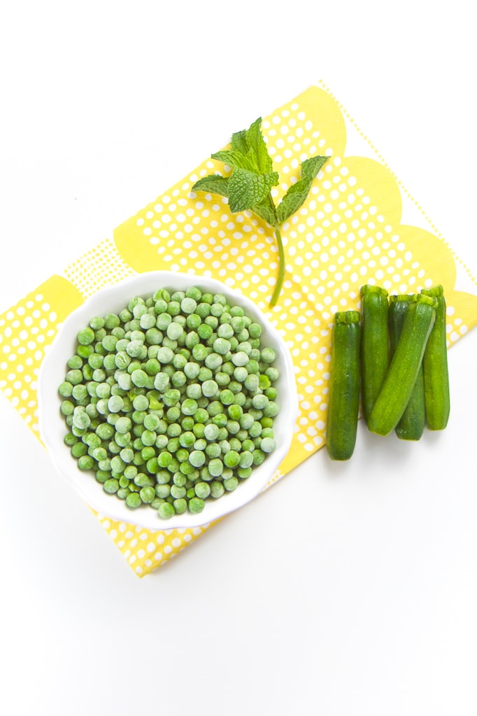 Peas in a white bowl, mini zucchini and mint sitting on top of a yellow napkin. 