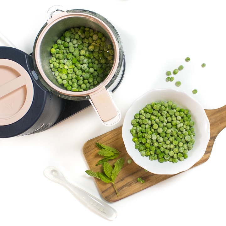 Spread of a black and pink BEABA babycook with a cutting board with peas and mint on it. 