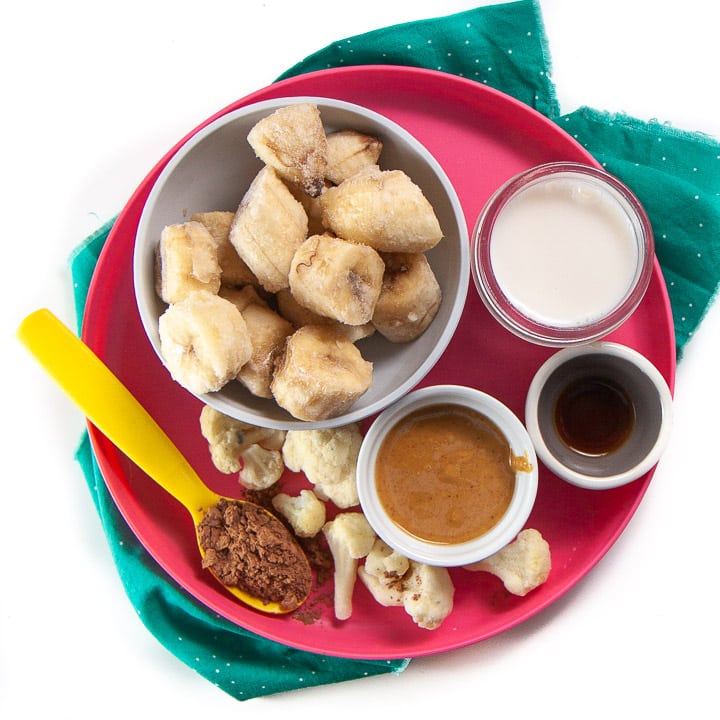 Plate full of smoothie ingredients for toddlers and kids - banana, peanut butter, chocolate, cauliflower, vanilla and milk. 