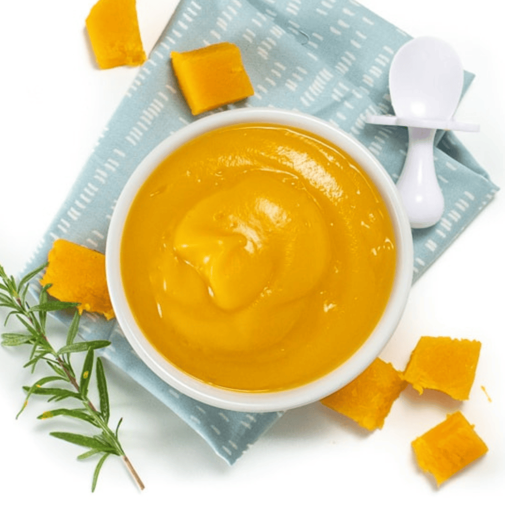 White bowl of smooth butternut squash puree on a blue napkin sitting on my white kitchen counter. With a white spoon and chunks of butternut squash and rosemary.