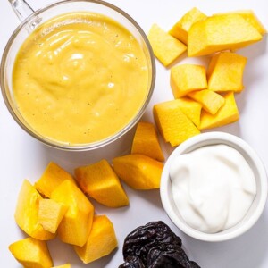 Image is of a white cutting board with produce scattered with a small bowl filled with baby food puree.