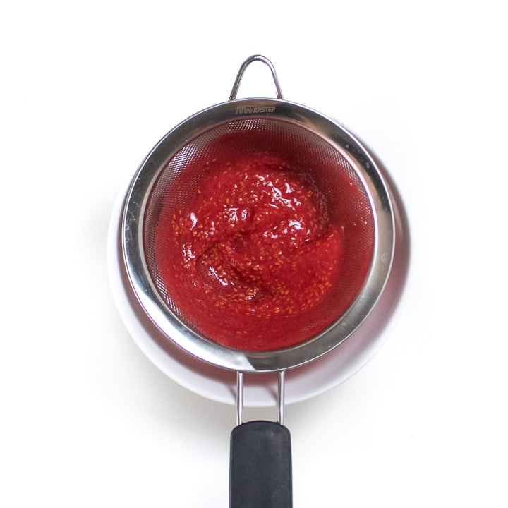 Fine mesh colander resting on a white bowl with raspberry seeds in it.