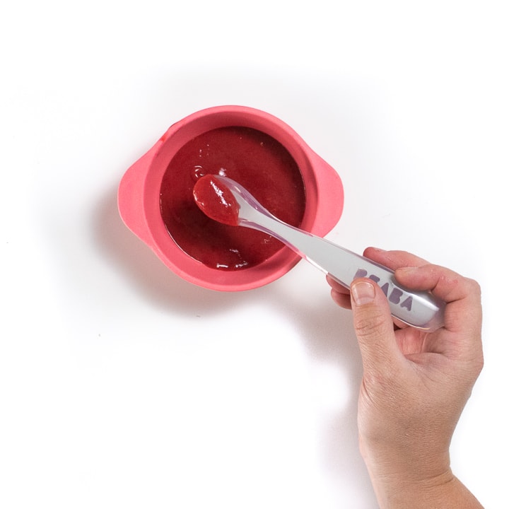 Pink bowl with hand holding a gray spoon with raspberry puree in it.