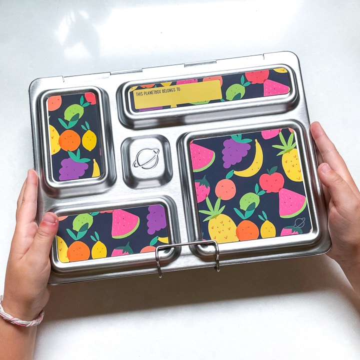 Small kids hands holding a closed planetbox lunch box with fruit magnets on the front.