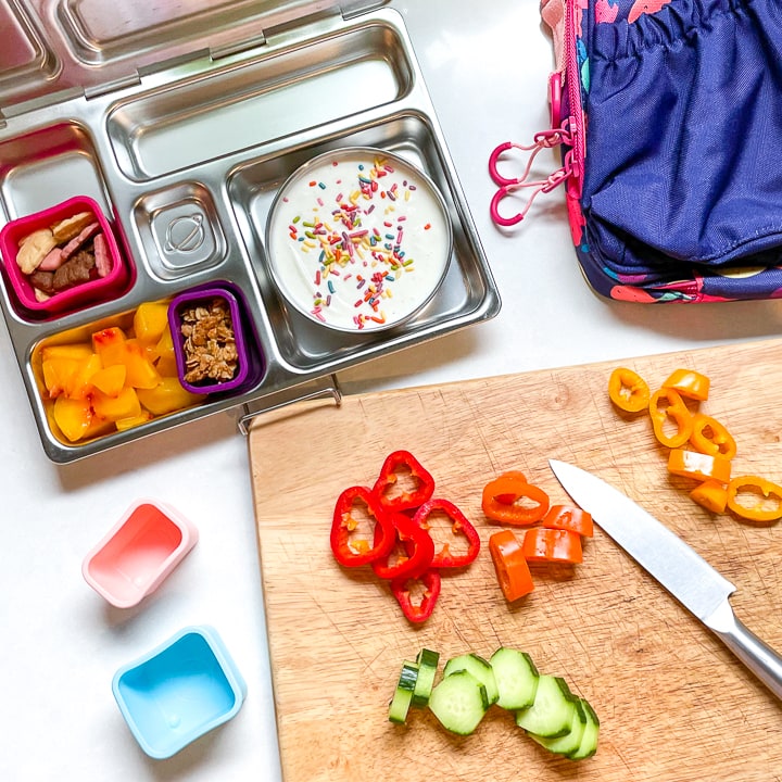 Cutting board with veggies and a planetbox lunch box sitting next to it halfway full with a kids lunch.