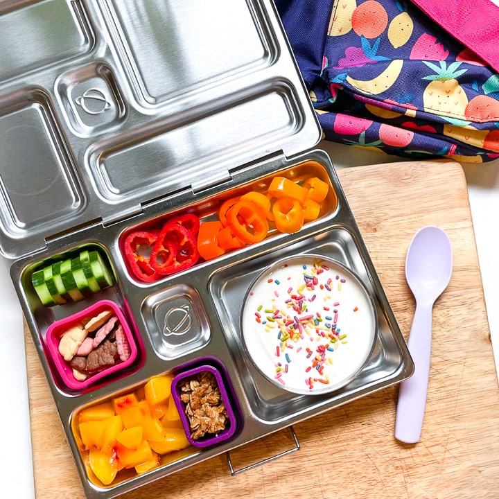 Planetbox on a cutting board filled with healthy and colorful food for a kids lunch with a purple spoon rising next to it.