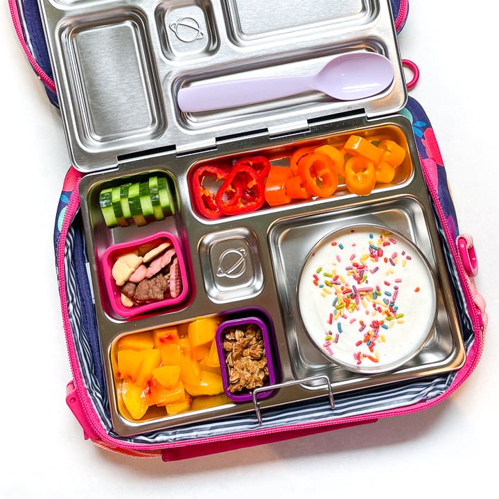 Planetbox lunch box filled with a colorful lunch is sitting inside a lunch box with a purple spoon resting on the lid.