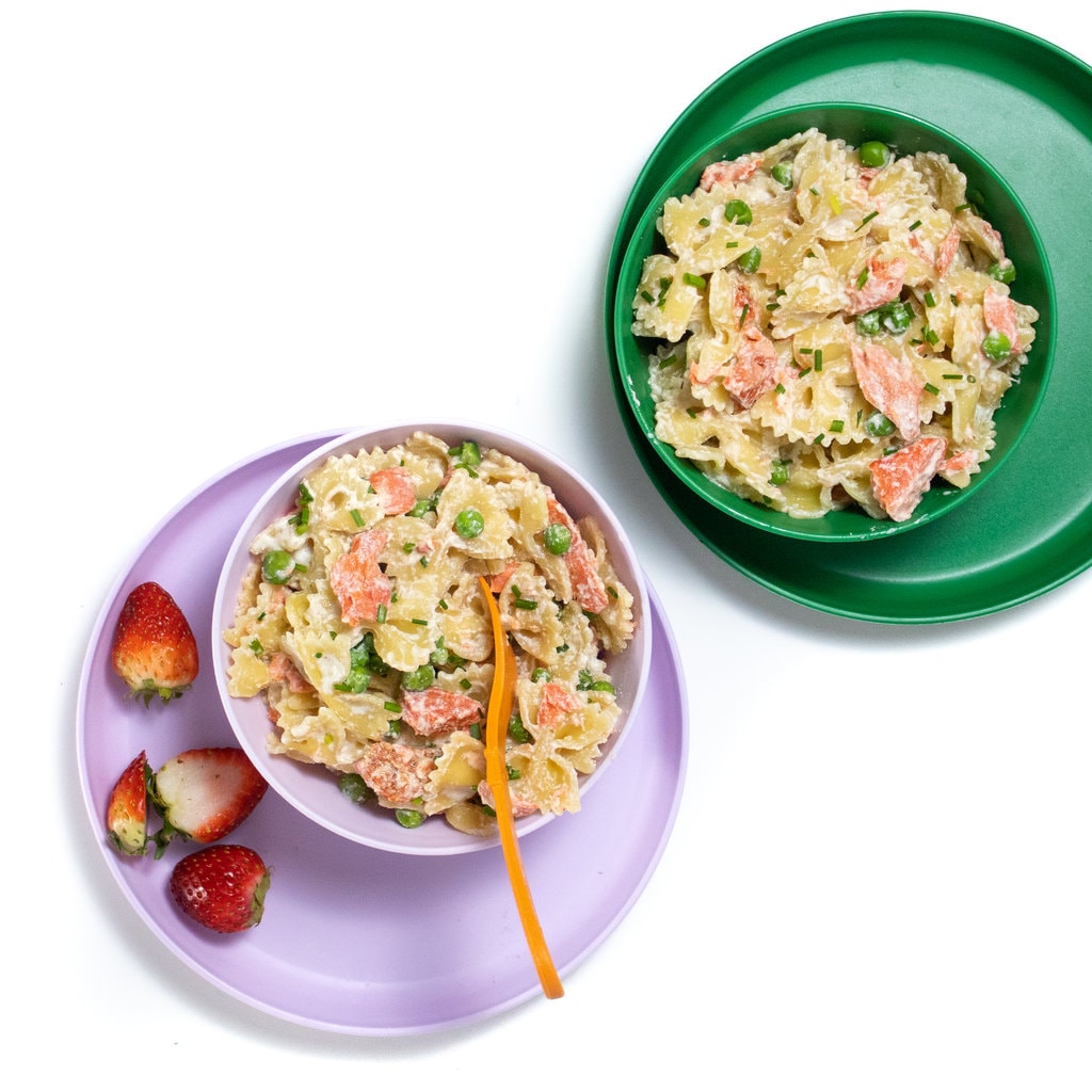 Set of purple and green kids bowls and plates with salmon pasta with peas inside.