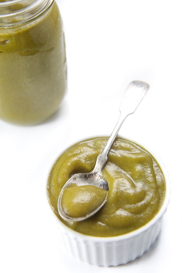 Small white bowl with spinach apple puree inside with baby spoon resting on top with jar of puree in the background.