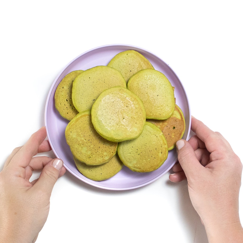 Two hands holding a baby plate with a stack of spinach banana pancakes.