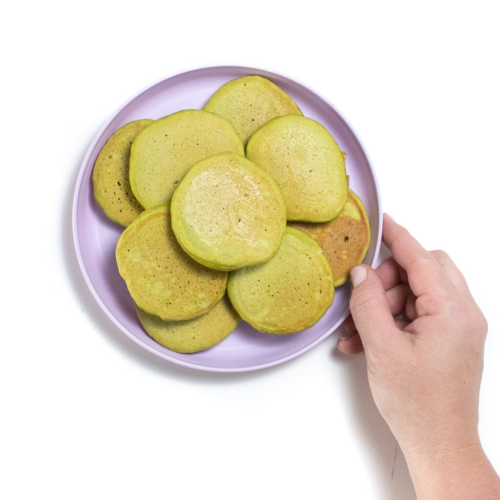 A purple toddler play with a stack of spinach pancakes with a hand holding the plate.