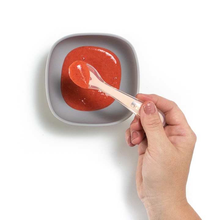 Hand holding a pink spoon with strawberry puree in it that is hovering over a bowl of strawberry puree.