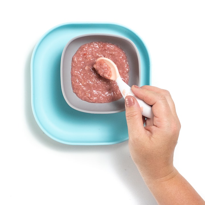 Hand holding a white spoon stirring a strawberry oatmeal inside a gray bowl.