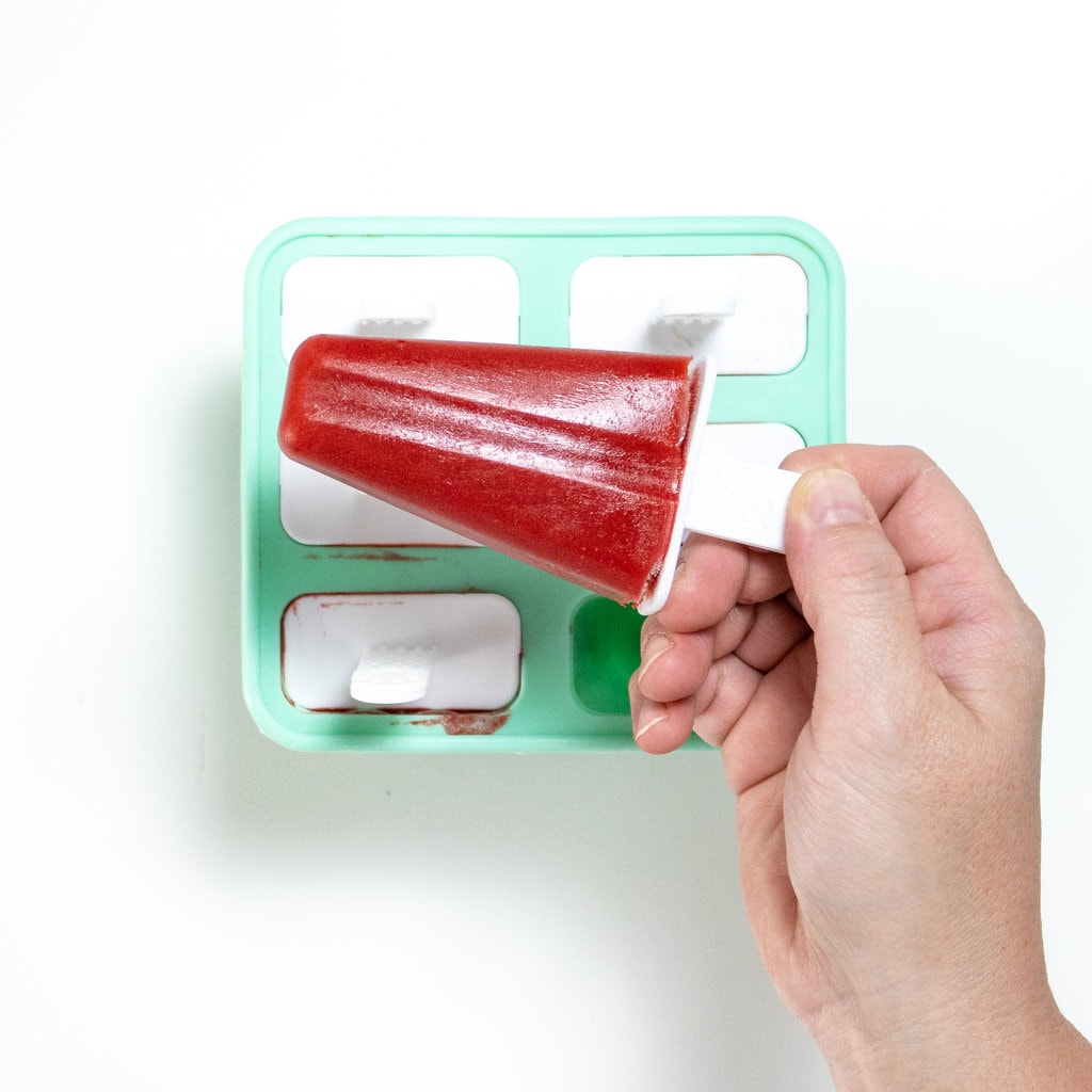 A hand holding a bright pink popsicle over a popsicle tray I guess the way background.