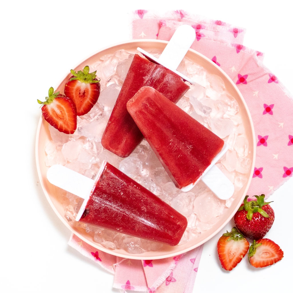 A pink kids plate with ice cubes and three strawberry popsicles, with strawberries on the side in a pink napkin against a white background.