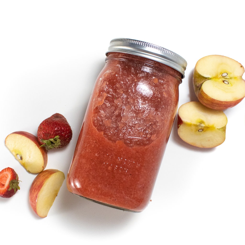 A class jar with strawberry applesauce with strawberries and apples scattered around it against away background.