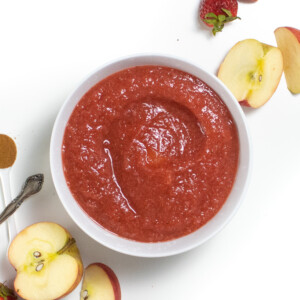 A white bowl against a white background with chopped apples and strawberries scattered around it.