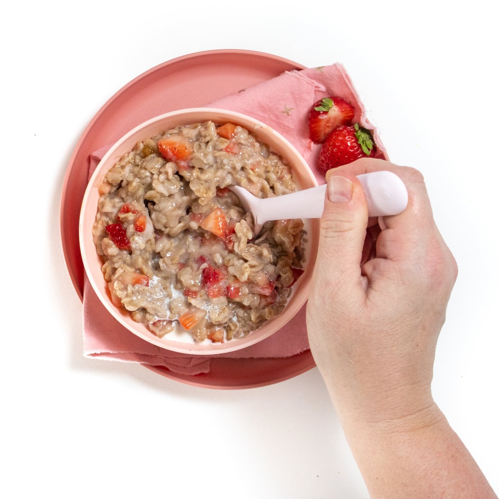 Kids, pink bowl and plate filled with strawberry oatmeal with hands during the oatmeal with a pink spoon.