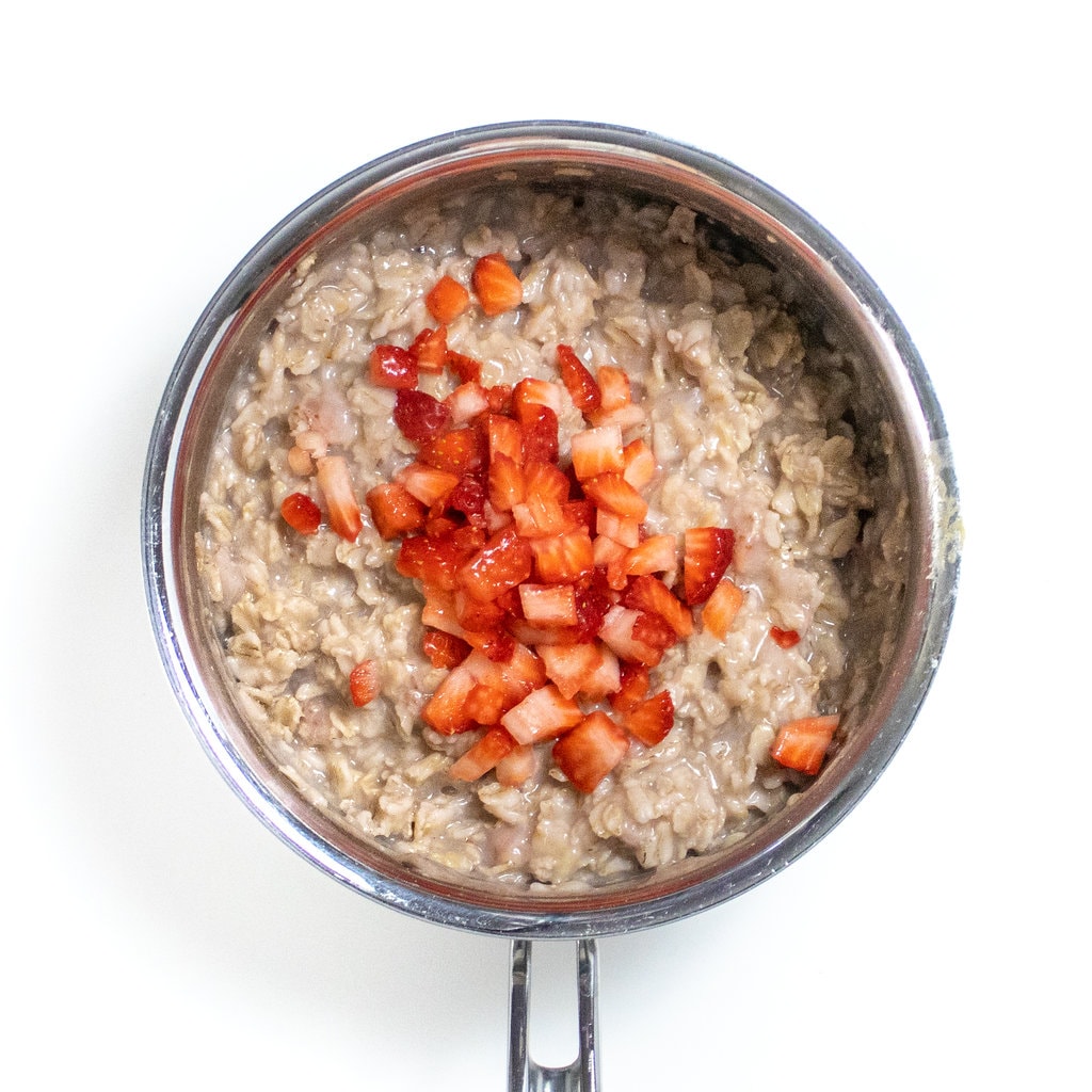 Silver sauce pan with cooked strawberry oatmeal with more finely chopped strawberries on top.