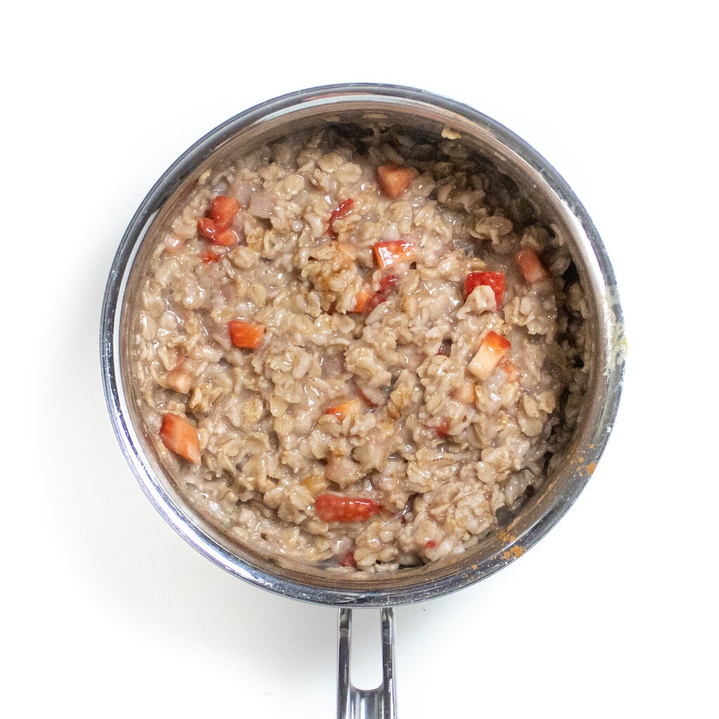 A small silver sauce pan with cooked strawberry oatmeal with mashed bananas on a white countertop.