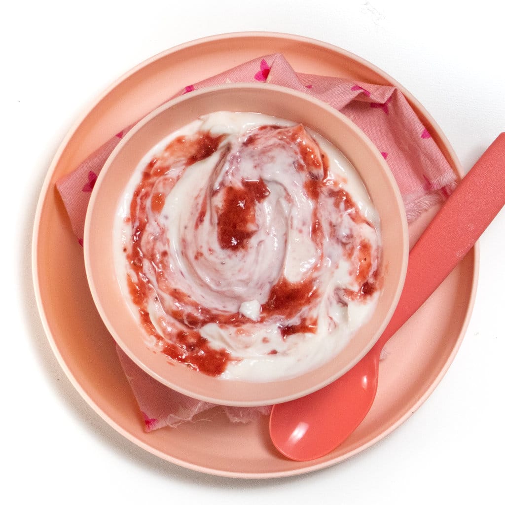 A pink bowl and plate filled with a white yogurt and strawberry purée swirled in against a white background.