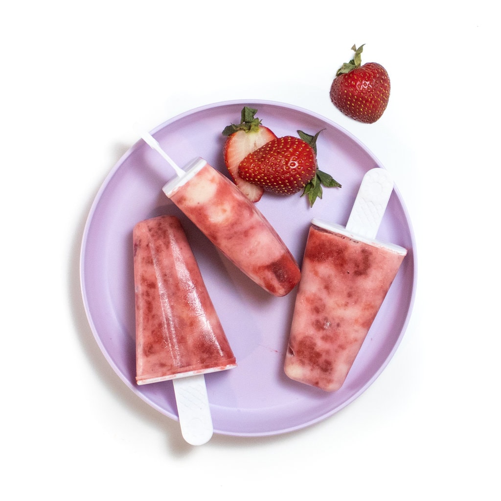 03 strawberry yogurt popsicles with sliced strawberries on a purple plate against the way background.