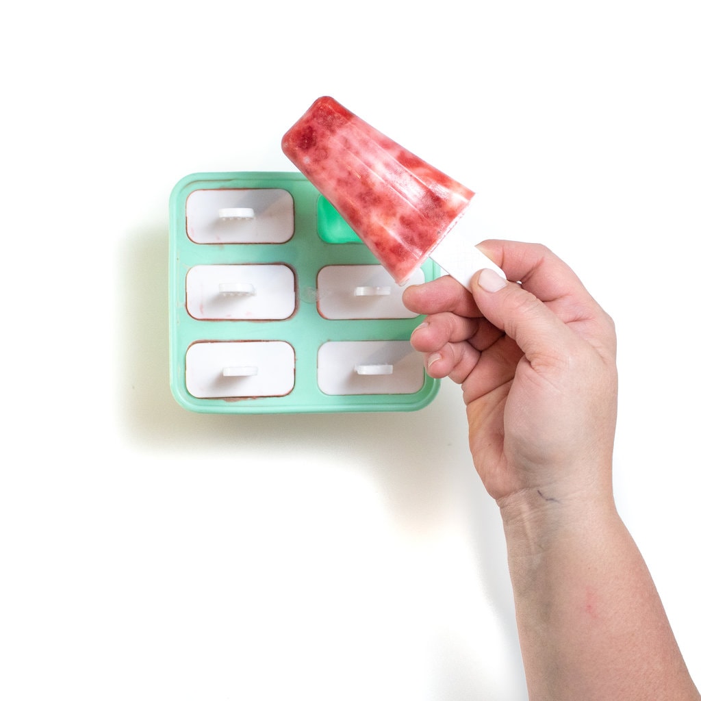 A teal popsicle tray with the hand holding a strawberry yogurt popsicle on top of it.