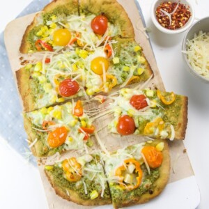 Whole wheat veggie pizza on a cutting board with side bowls of parmesan cheese and red pepper flakes.