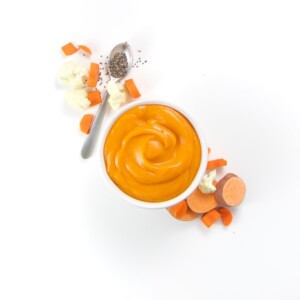 A small white bowl filled with creamy nutrient rich homemade baby food puree with produce around it.