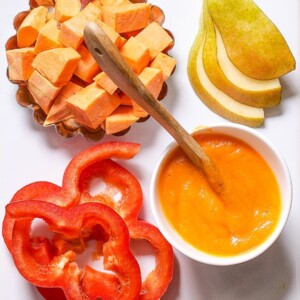 White cutting board with a spread of produce on it with a bowl of smooth baby food.