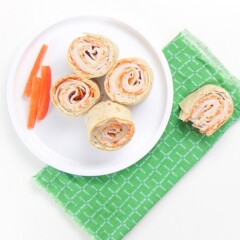 a small white plate sitting on a green napkin on top of a white surface. Place has 4 turkey and carrot rollups wit 3 slices of red peppers on the plate. On the napkin is one half eaten rollup.