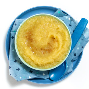 Blue kids bowl on a blue plate and blue napkin filled with homemade applesauce with spoon resting on top and a sprinkle of cinnamon on top of the applesauce.