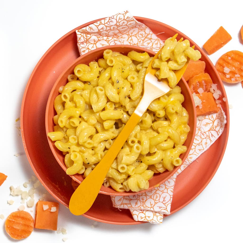 A pink bowl on top of the pink plate with a colorful napkin with veggies scattered on the white countertop. The bowl is full of veggie mac & cheese.