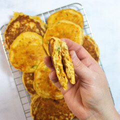 My hands holding a torn half veggie pancake to show the veggies inside over a wire rack of veggie pancake on my kitchen counter.