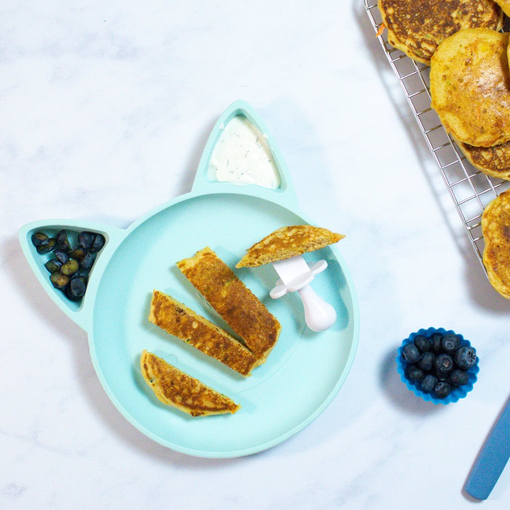 A toddler bear plate with veggie pancakes, a side of cut, blueberries and a side of ranch with the rest of the ingredients scattered around.