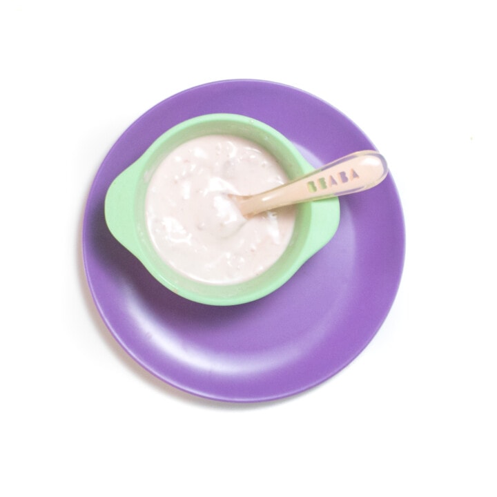 A peach baby spoon resting on a teal baby bowl with a chunky watermelon and yogurt purée that is sitting on a purple plate against the white back out.