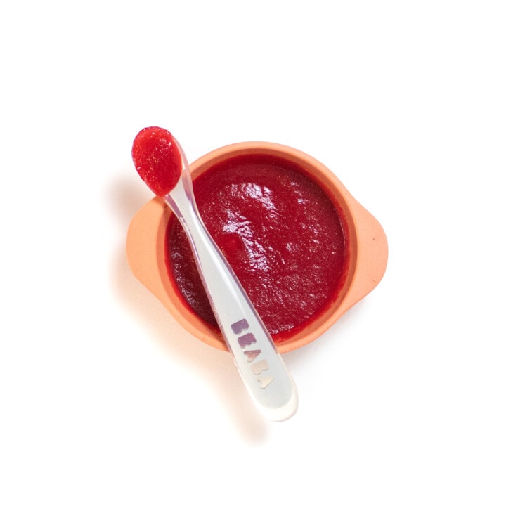 A peach baby bowl with a gray baby spoon resting on top holding a bright pink purée against away background.